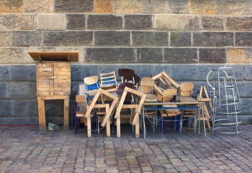A collection of discarded furniture and wooden items arranged along a cobbled pavement in front of a weathered brick wall. The items include various wooden chairs, some with a natural finish and others painted, stacked and leaning against each other in a disorderly manner. There is a small wooden cabinet with a slotted front on the left, and a silver metal trolly frame on the right. The background features a grey, aged brick wall with visible texture and mortar lines, suggesting an outdoor urban setting suitable for rubbish removal services in Mayfair, London. The scene appears to depict waste awaiting collection, with the furniture and wooden debris positioned on the pavement, ready for disposal or pick-up by a specialist such as Commercial Waste Removal Mayfair.
