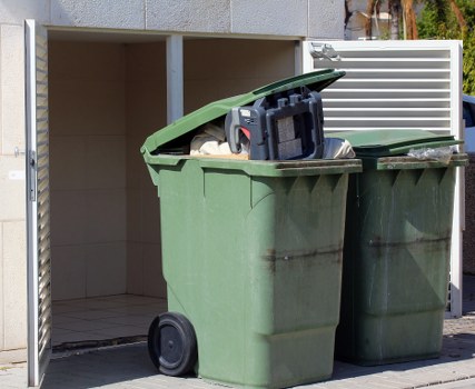 An outdoor scene showing a green wheeled wheelie bin with its black lid open, revealing various waste items inside, positioned on a paved driveway next to a small, partially enclosed structure with beige tiled flooring and a white slatted privacy screen. The bin's surface appears slightly weathered with streaks of dirt or stains, and the surrounding environment suggests a residential or commercial property in Mayfair. The image demonstrates typical rubbish collection equipment used by commercial waste removal services, with the bin placed ready for disposal or collection, set against a backdrop of outdoor fixtures and urban landscaping.