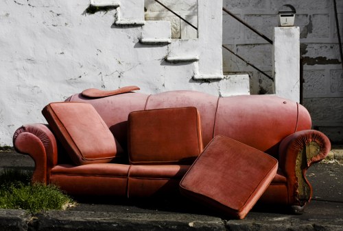 A large, reddish-brown fabric sofa with ornate rolled arms and carved wooden details on the armrests, positioned outdoors on a concrete surface in front of a weathered white brick staircase with metal railing. The sofa has two matching cushions and an attached armrest on the left side, while a matching rectangular ottoman, also upholstered in the same fabric, is placed at an angle on the right side, partially leaning against the sofa. The setting appears to be a yard or alleyway area near a residential or commercial building, with some patches of grass and dirt visible around the sofa, suggesting it has been discarded as rubbish. The overall scene reflects a typical example of bulky waste awaiting removal, relevant to rubbish clearance services in the Mayfair area.