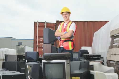 A young male worker wearing a yellow safety helmet and a high-visibility vest stands outdoors amidst a collection of discarded electronic devices, including old monitors, computer towers, and other obsolete technology. The objects are placed on a paved surface, with some stacked or leaning against each other, and appear to be in various stages of disarray, suggesting clearance or disposal of electronic waste. In the background, a red shipping container and industrial warehouse buildings are visible under a cloudy sky, indicating an industrial or commercial area likely within or near Mayfair, London. The scene reflects a scenario of electronic waste collection, fitting within the scope of rubbish removal services provided by Commercial Waste Removal Mayfair, and highlights the importance of proper disposal of electronic items in compliance with environmental regulations.