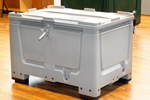 A large, industrial-grade gray plastic waste bin with a secure, hinged lid and a metal latch, positioned on a polished wooden floor in an indoor setting. The bin features reinforced edges and sturdy corner supports, indicating durability for heavy-duty rubbish collection. It is situated near a green waste container and other bins, with additional waste management equipment partially visible in the background. The environment appears to be a storage or waste collection area, with lighting providing clear visibility of the textured surface and color tones of the bin. The scene reflects a professional setup for rubbish removal services, relevant to commercial waste management in Mayfair or similar urban areas. Commercial Waste Removal Mayfair, specializing in rubbish disposal, would utilize such containers for efficient waste handling and collection.