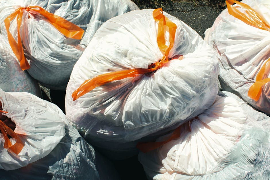 A close-up view of several large white plastic rubbish bags filled with waste materials, secured with orange plastic ties. The bags are piled together outdoors on a paved surface, with some showing sl